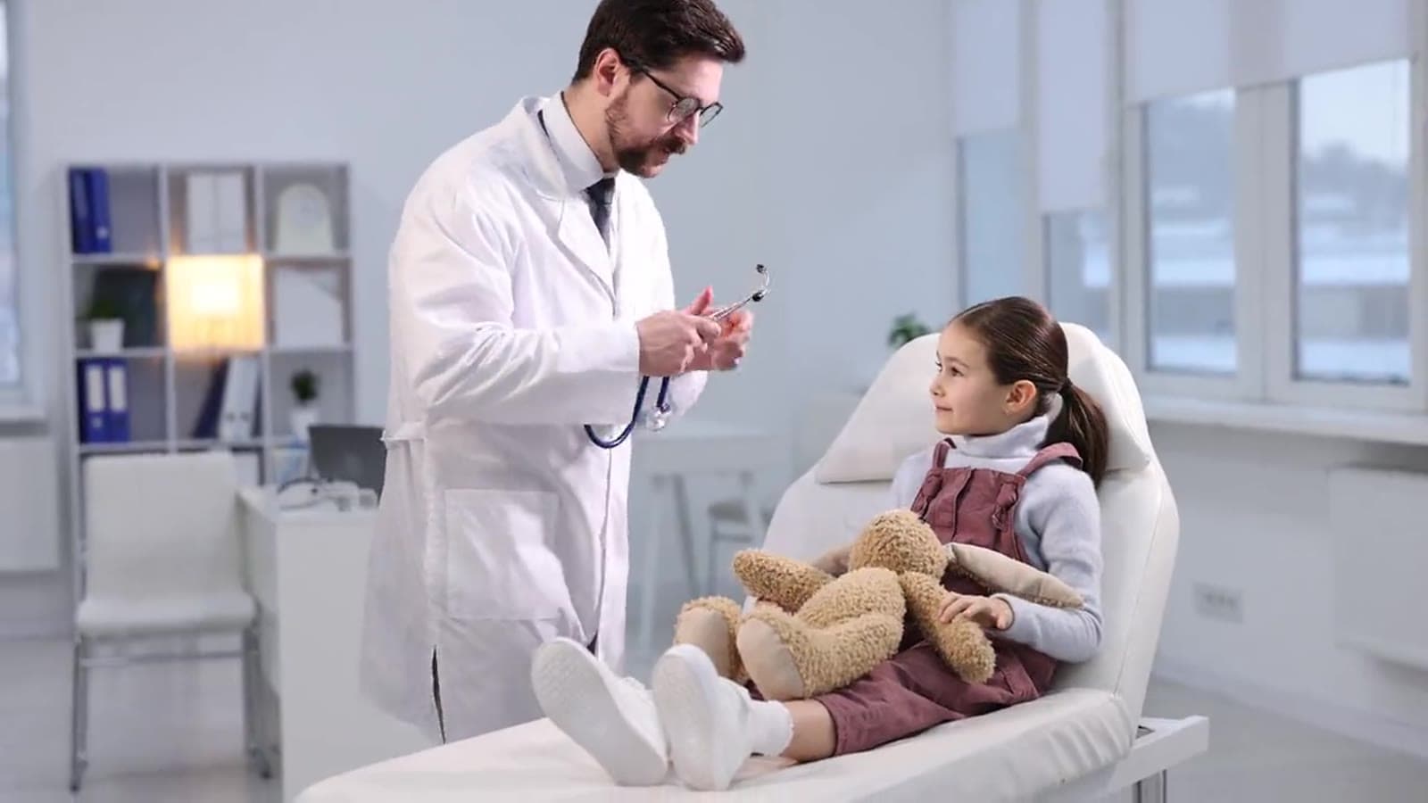 A pediatrician listens to a young girl’s heart with a stethoscope while she sits on the exam table holding a teddy bear
