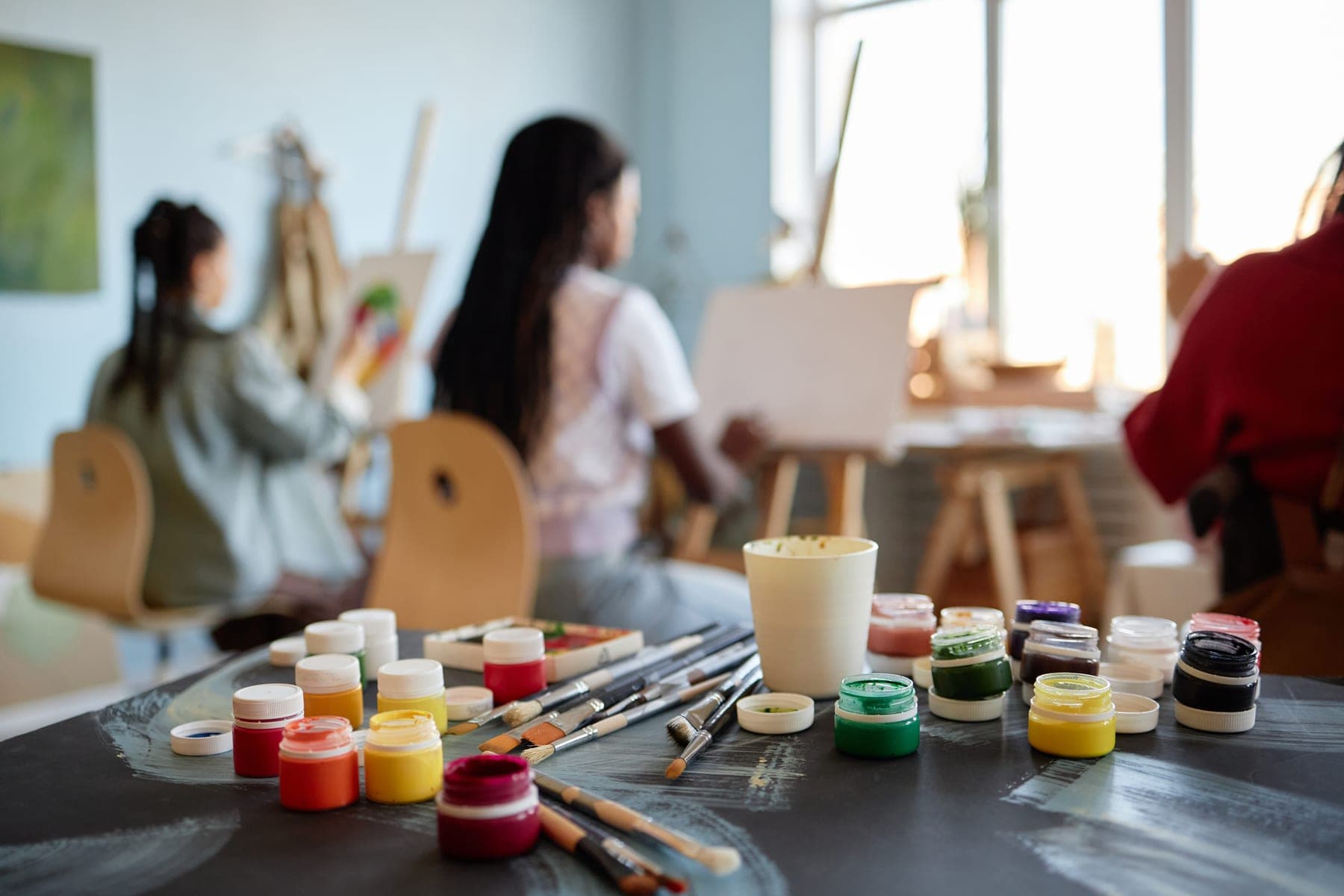 Jars of paint and brushes on a table in a bright art class, with painters at easels in the background