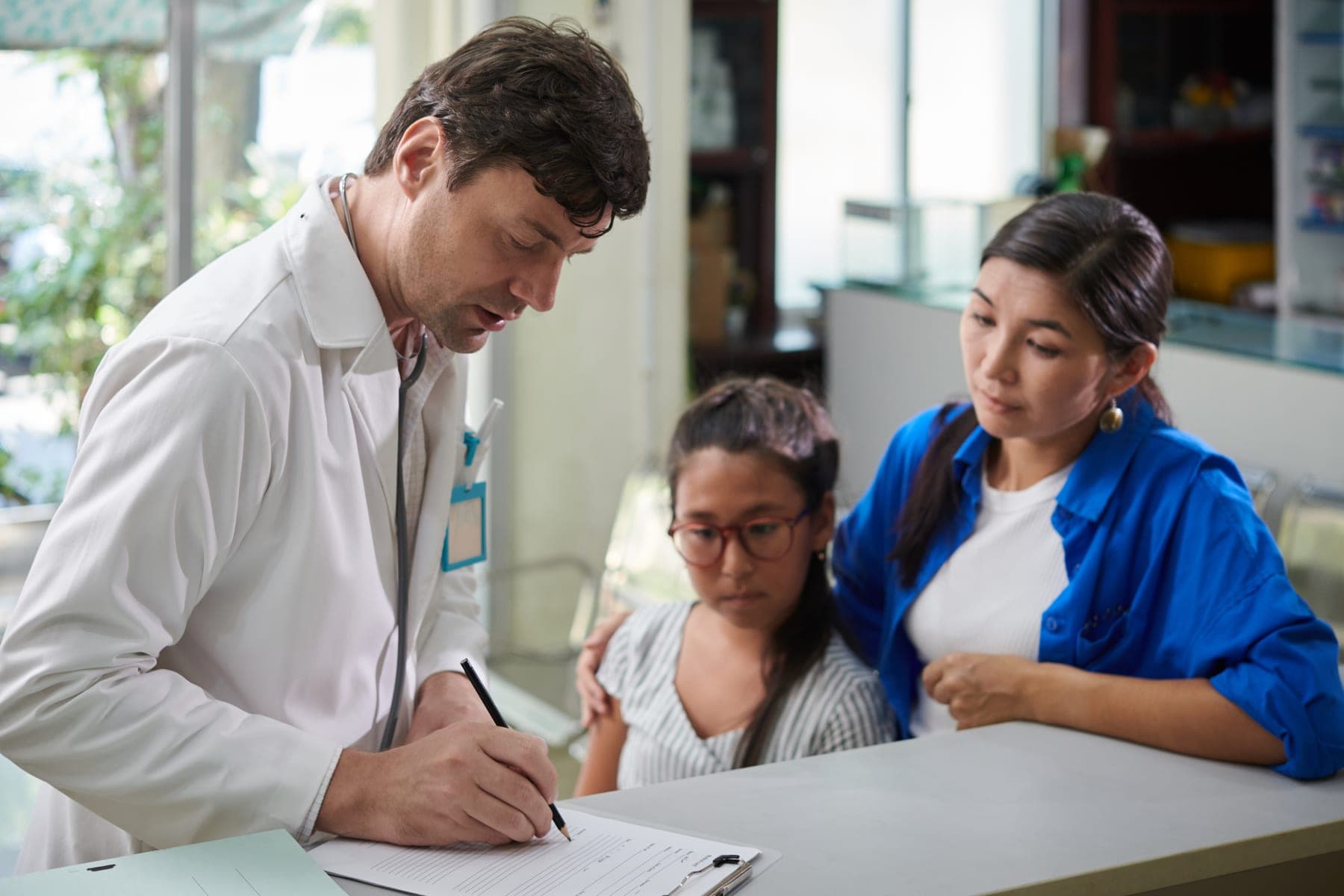 A doctor fills in a medical form at his desk while a mother and her daughter sit beside him