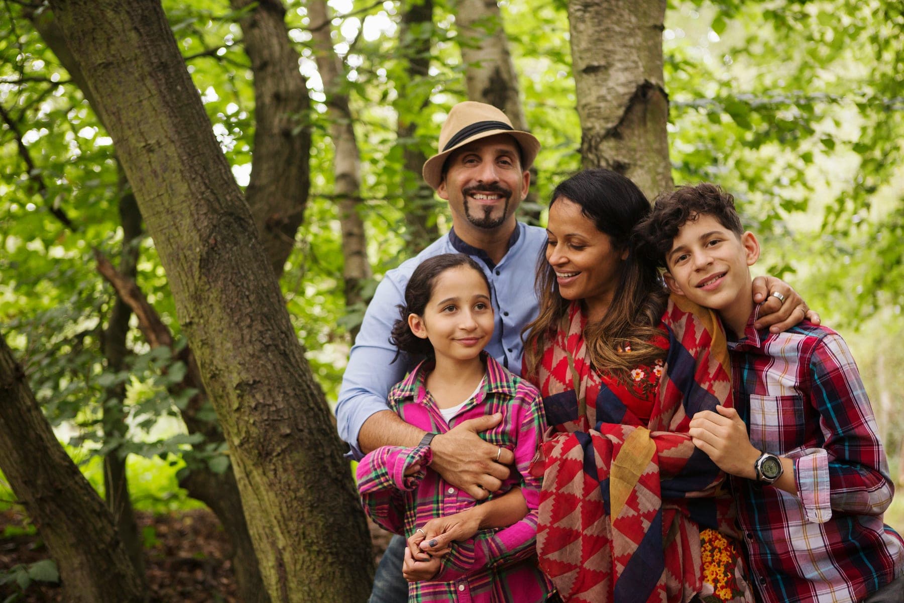 A smiling family of four stands together among the trees in a sunny forest