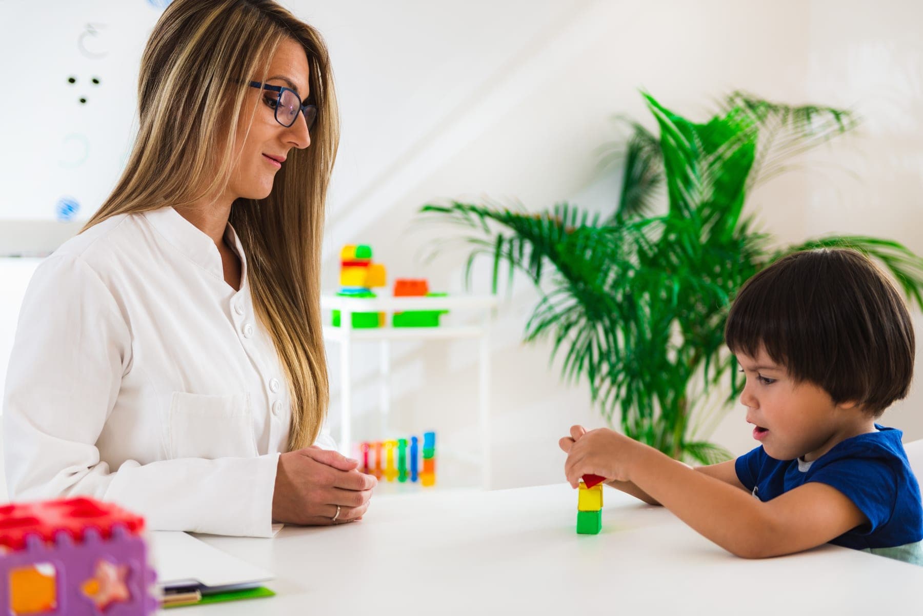A young boy stacks coloured blocks at a white table while his therapist watches