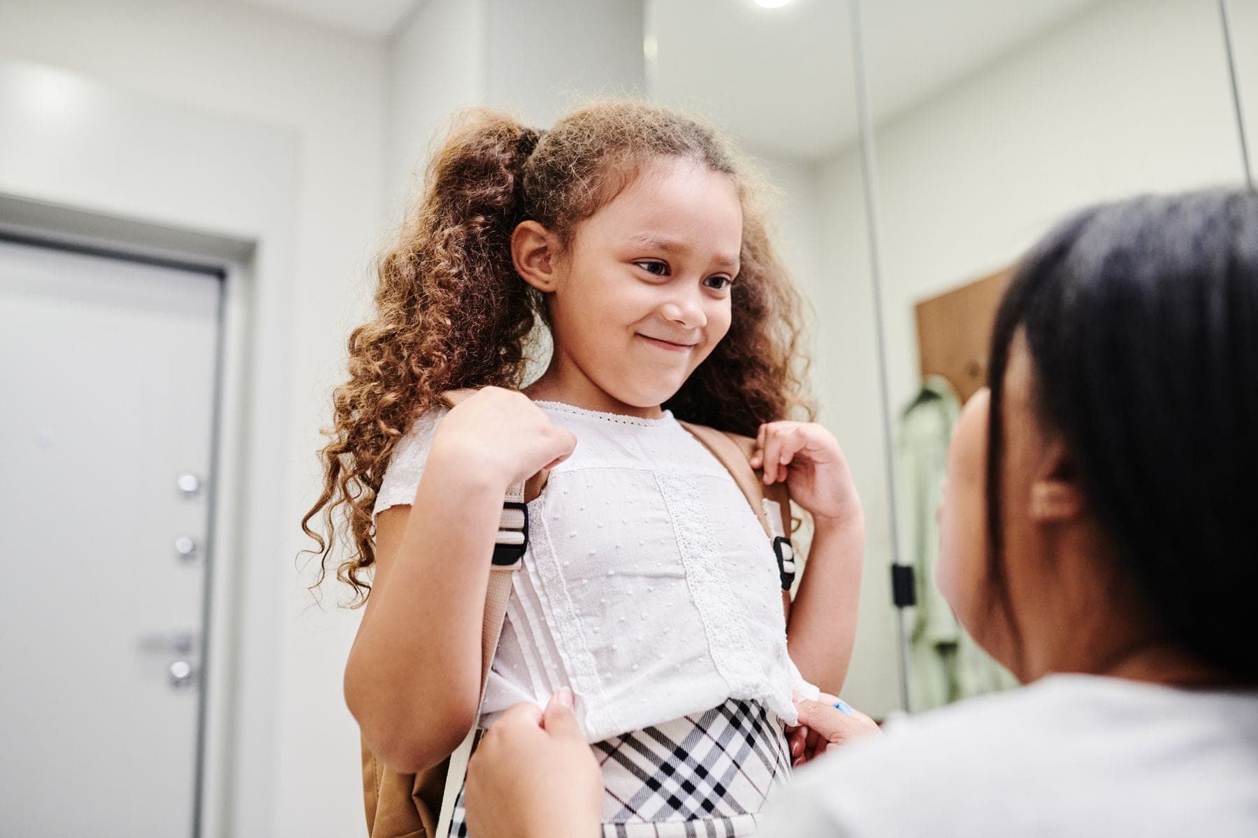 A smiling girl with curly hair puts on her backpack with her mother before school
