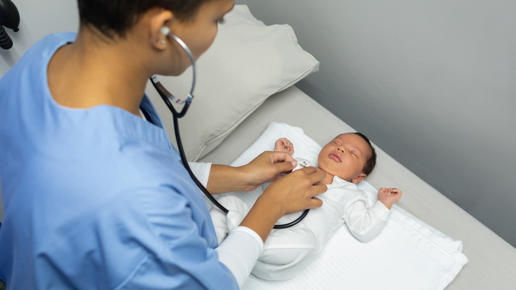 A nurse in scrubs listens to a newborn’s chest with a stethoscope in a hospital room