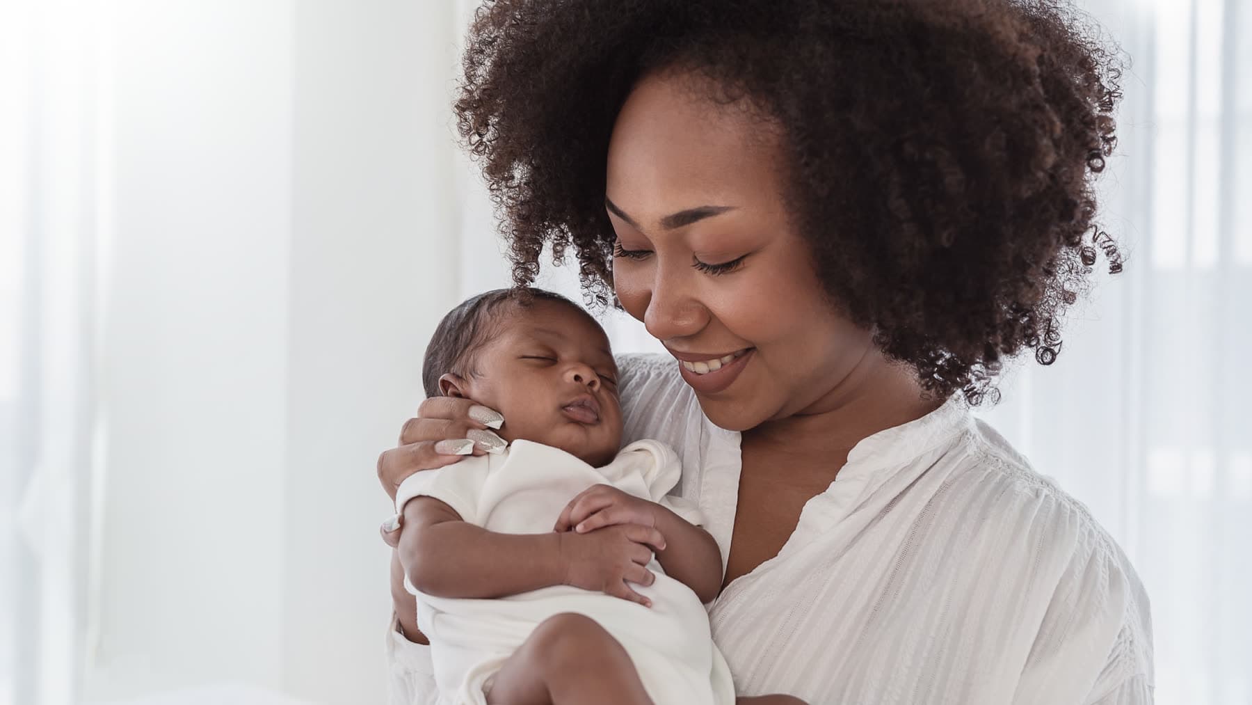 A mother holds her sleeping newborn against her shoulder by a bright window