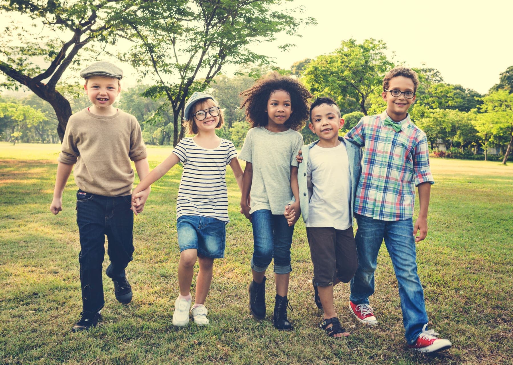 Five children of different backgrounds walk together across a sunny park, holding hands