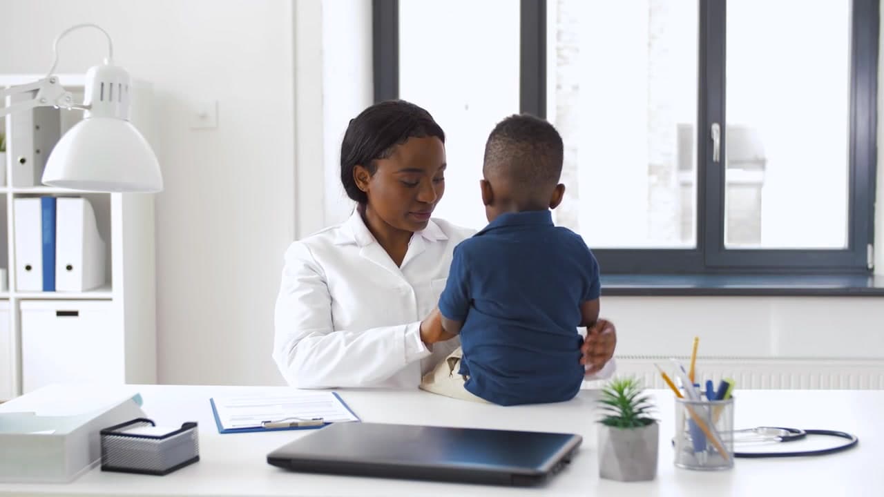 A pediatrician gently examines a young boy sitting on her desk in a bright office