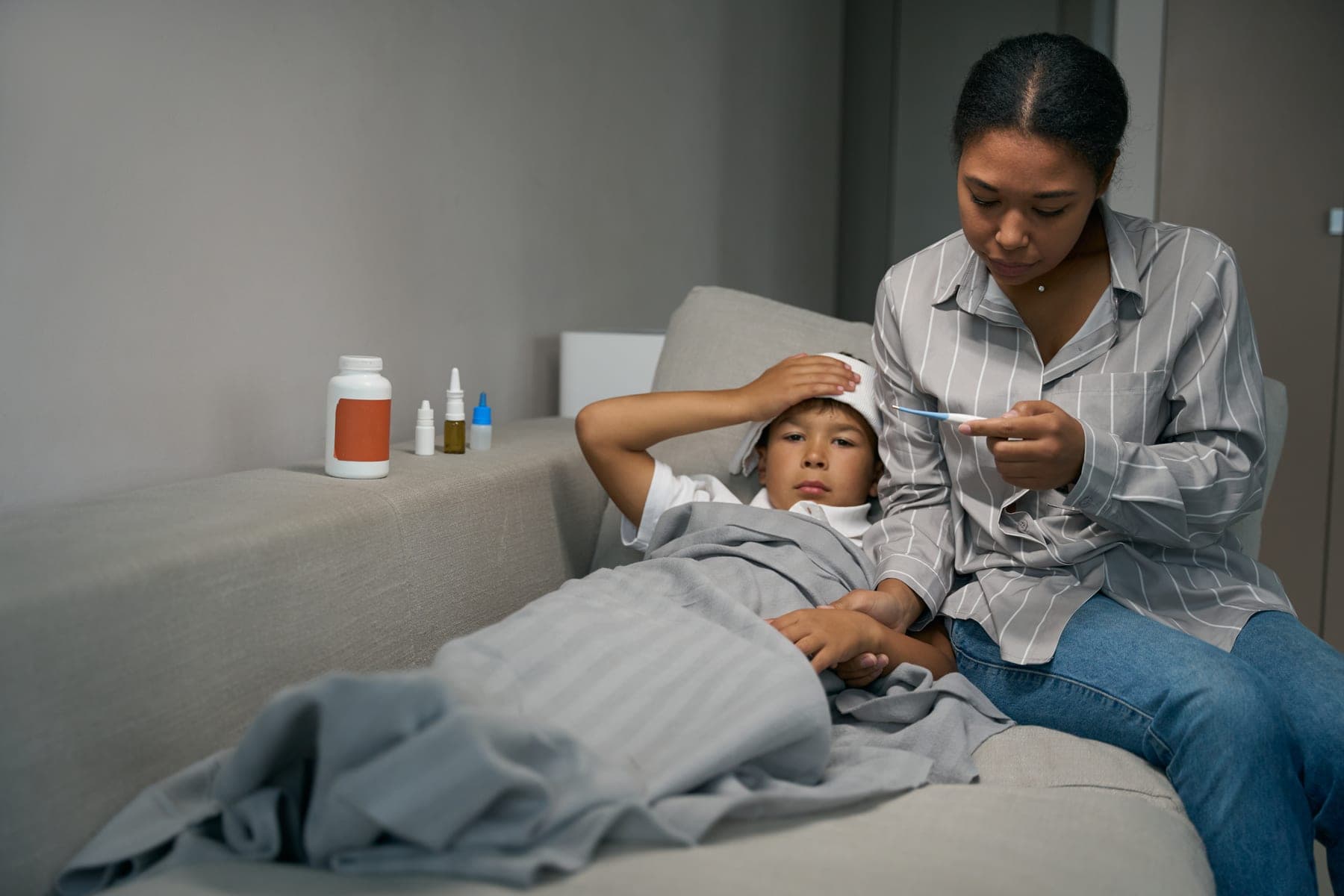 A mother checks her son’s temperature while he rests on the sofa with a cool cloth on his forehead