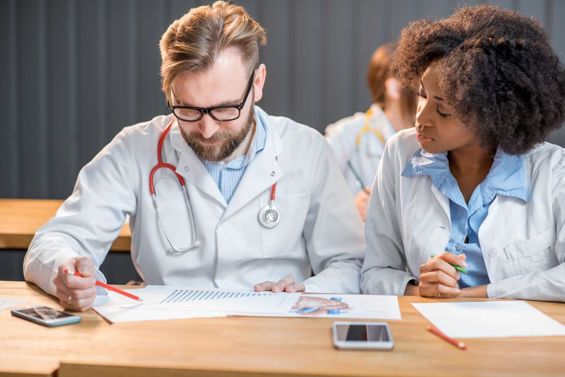 A physician and a student review patient charts together at a wooden table