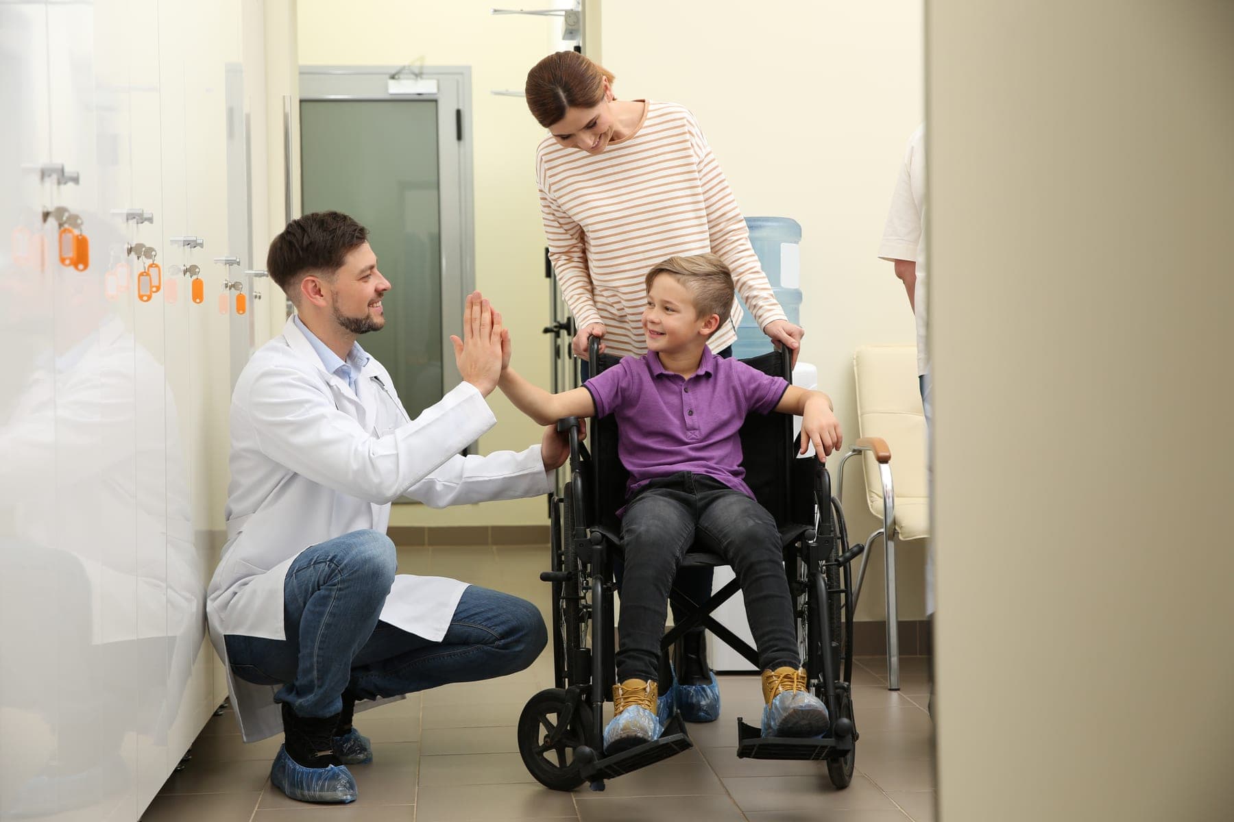 A pediatrician kneels to give a smiling boy in a wheelchair a high five while his mother looks on
